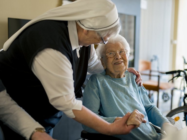 Eine Ordensschwester steht neben einer Frau, die im Rollstuhl sitzt und hat ihr einen Arm um die Schultern gelegt, mit der anderen Hand hält sie die Hand der Seniorin fest. Die Seniorin lächelt die Ordensschwester an.