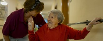 Haus St. Michael Eine ältere Frau im Rollstuhl hält sich an einer Stange an der Wand fest und unterhält sich lachend mit einer Pflegerin, die sich zu ihr hinunterbeugt.