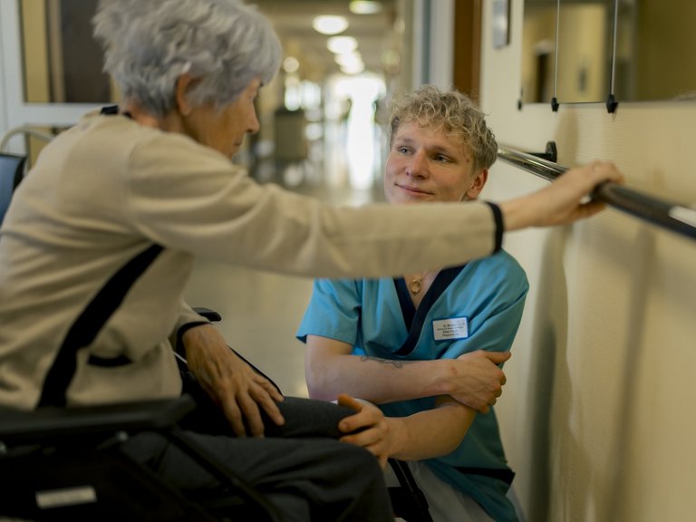 Ein junger Pfleger sitzt in der Hocke neben einer älteren Frau im Rollstuhl, die sich an einer Stange an der Wand festhält. Sie unterhalten sich.