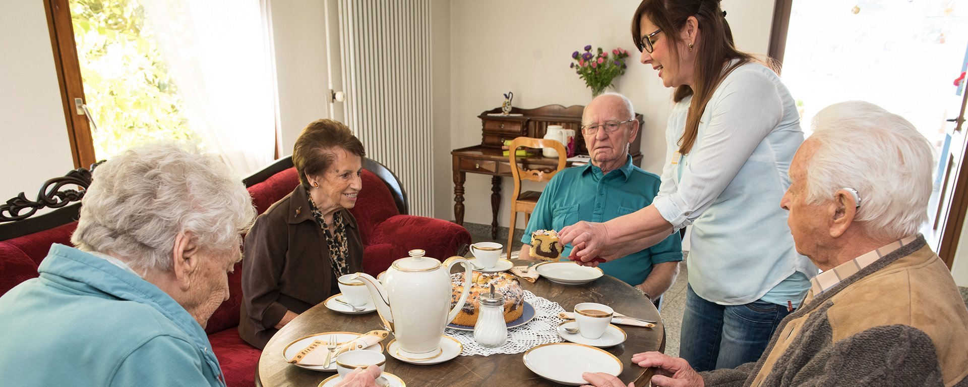 Bewohner beim Kaffetrinken an einem runden Tisch - eine Pflegerin schneidet und teilt Marmorkuchen aus
