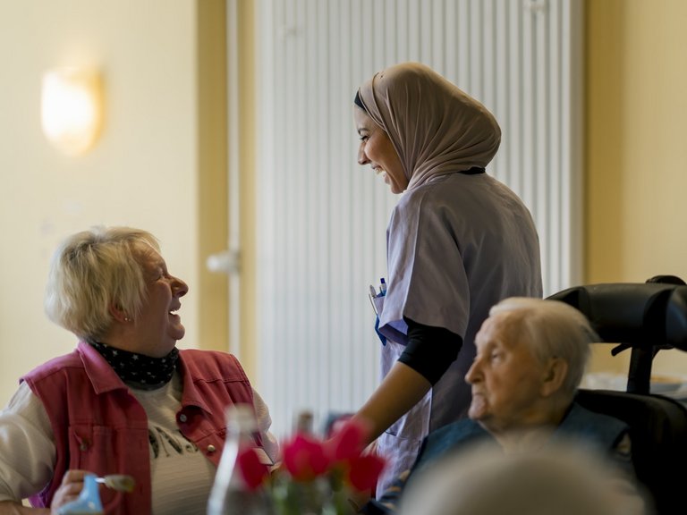 Zwei Pflegerinnen unterhalten sich, rechts daneben sitzt ein älterer Herr im Rollstuhl und beobachtet die Szene.