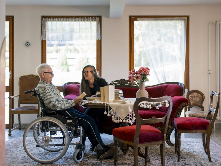 Eine Frau sitzt auf einer Couch neben einem Herrn im Rollstuhl, beide lachen.