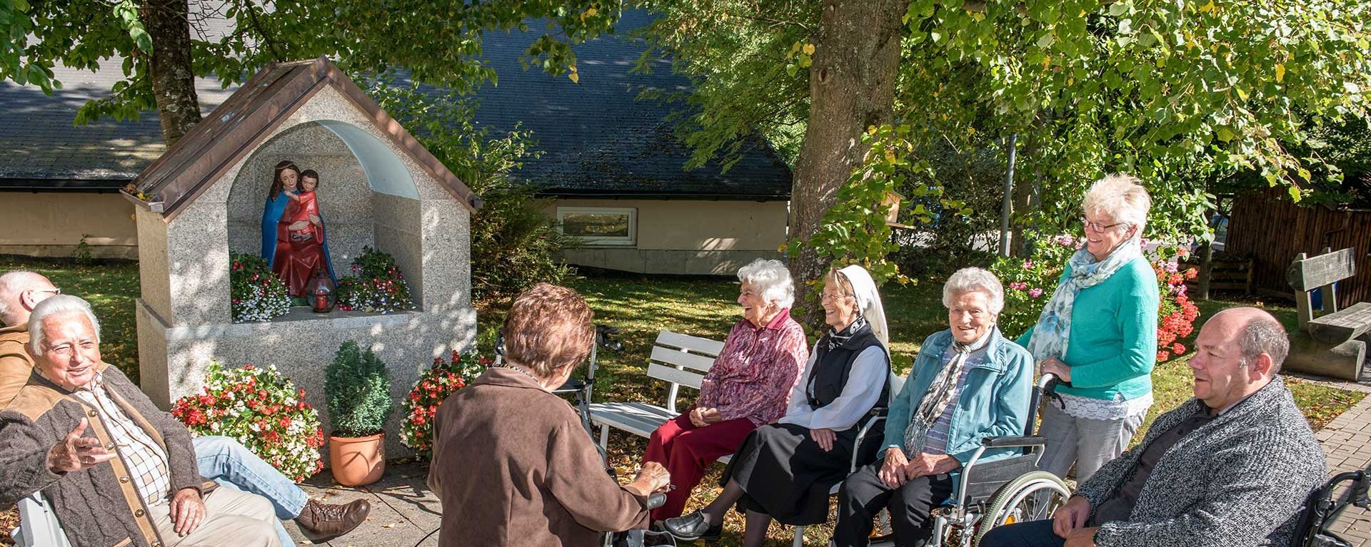 Eine Gruppe Bewohner unterhält sich vor einem Mariendenkmal auf Parkbänken